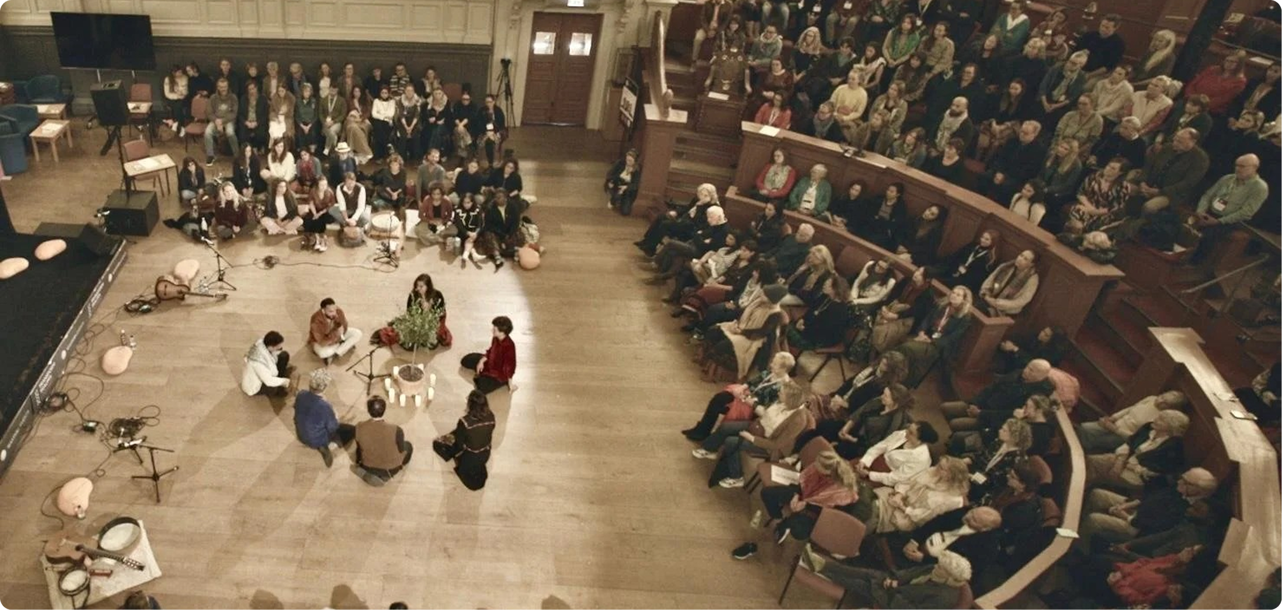Audience seated in a theater watching a stage performance or presentation, with musicians and a woman sitting on the floor in front of the stage, decorated with plants and candles.