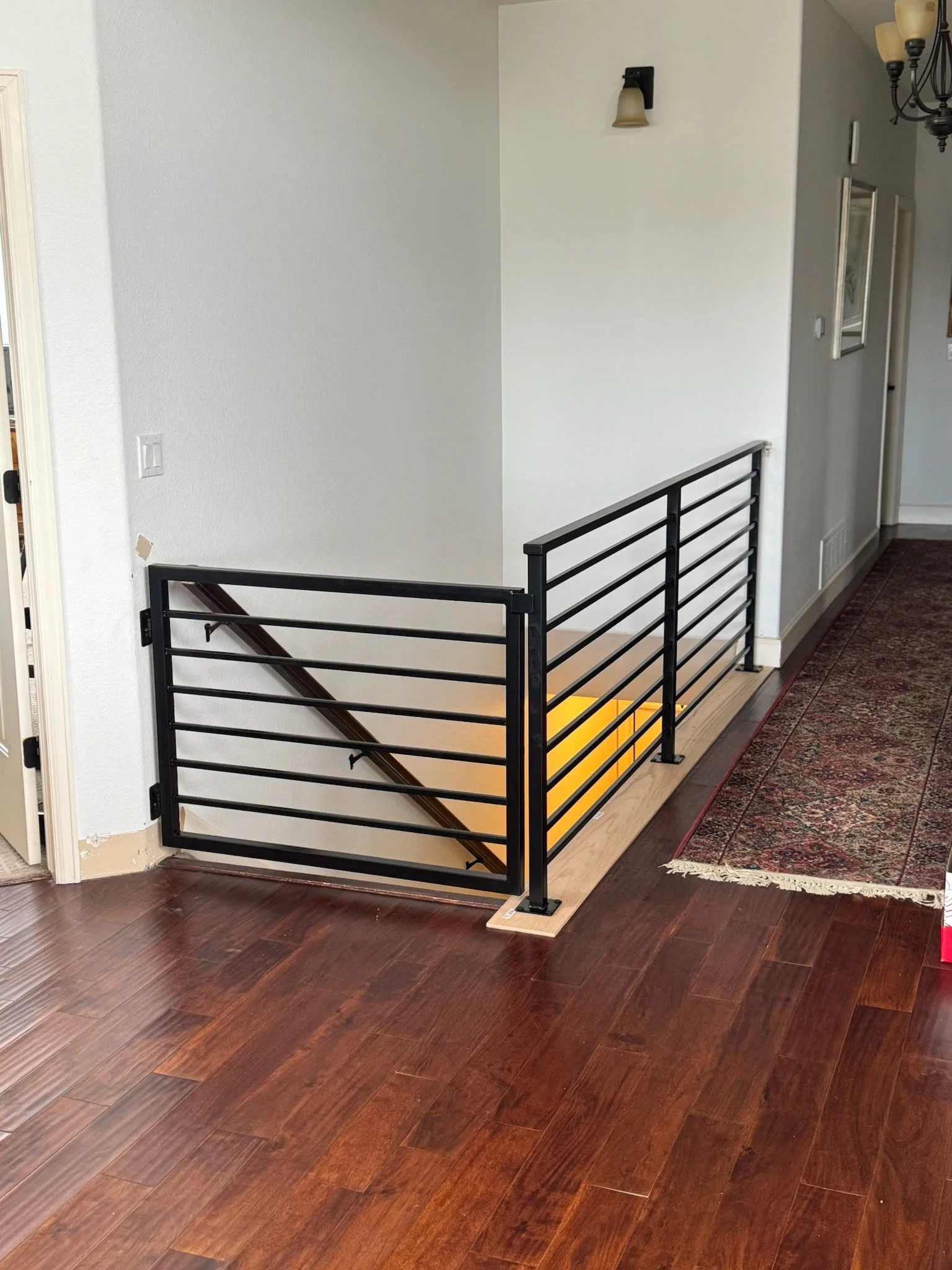 Interior view of a residential home with a staircase barrier; the barrier has a black metal railing with horizontal bars and a yellow stair tread behind it, situated on a wooden floor next to a hallway with a patterned area rug.
