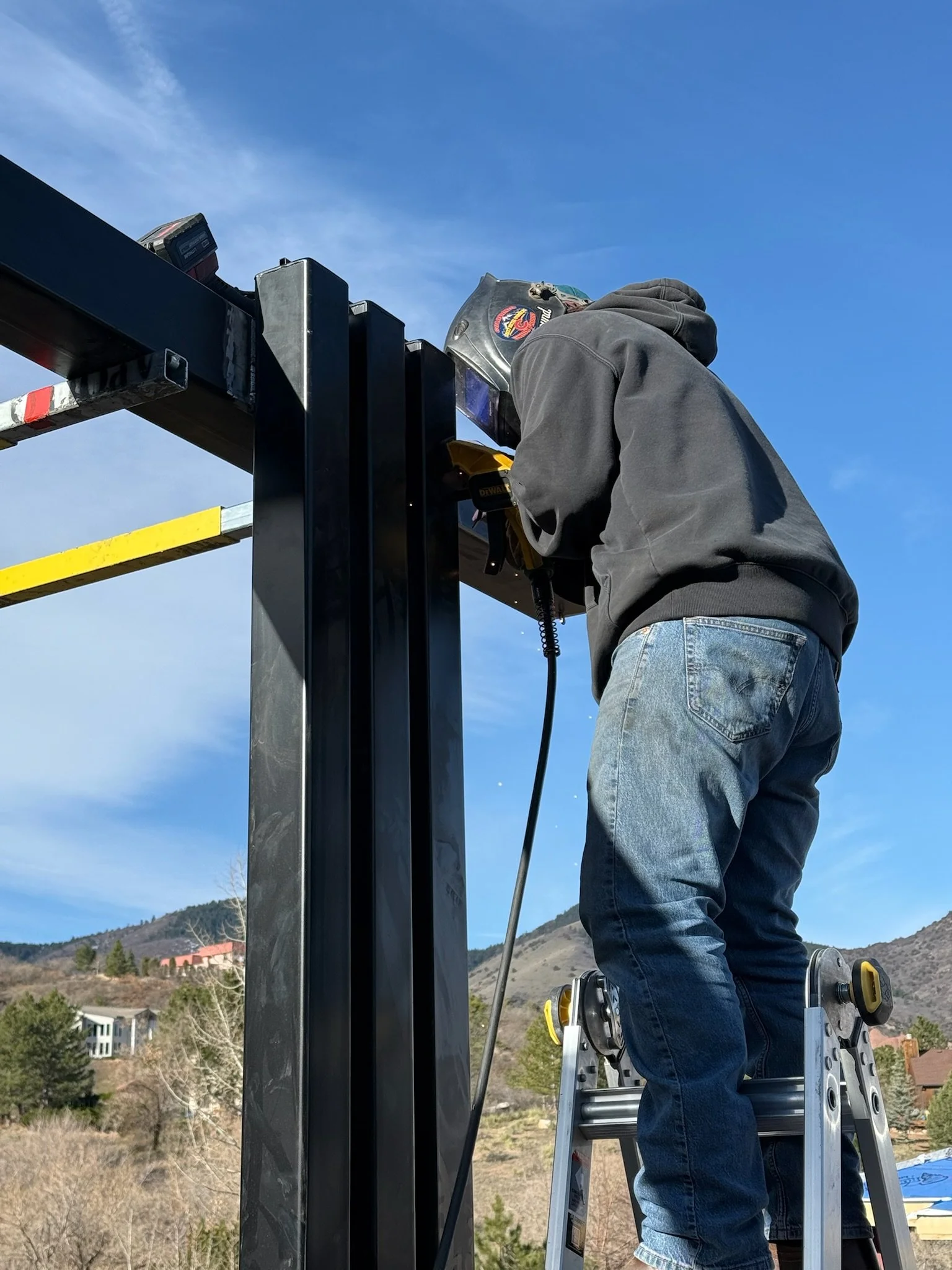 A worker wearing a welding helmet and hoodie working with tools on a steel structure outdoors with a mountainous landscape in the background.