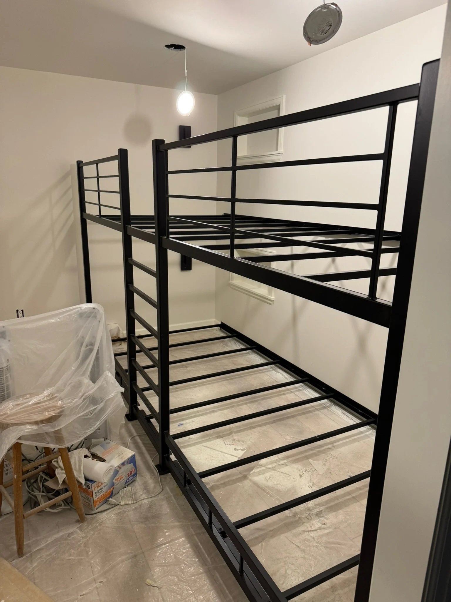 Black metal bunk bed frame in a room under construction or assembly, with a ladder attached, and a plastic-covered chair and box nearby.