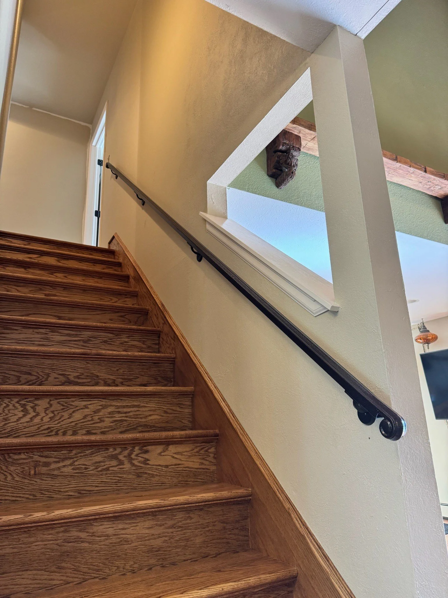 Wooden staircase with a dark handrail attached to a light-colored wall, overlooking an open space with a green accent wall and visible wooden beams in the ceiling.