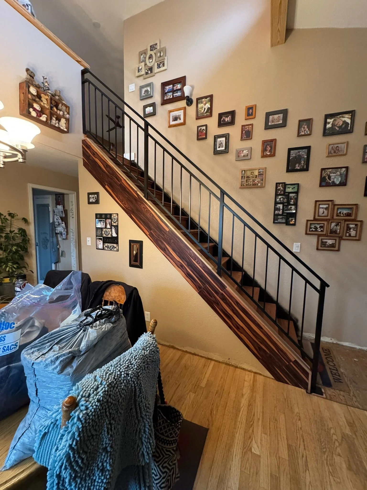 Interior of a house featuring a staircase with a black railing, a collection of picture frames on the beige wall, and a dining area with chairs, a table, and various items including bags and a blanket.
