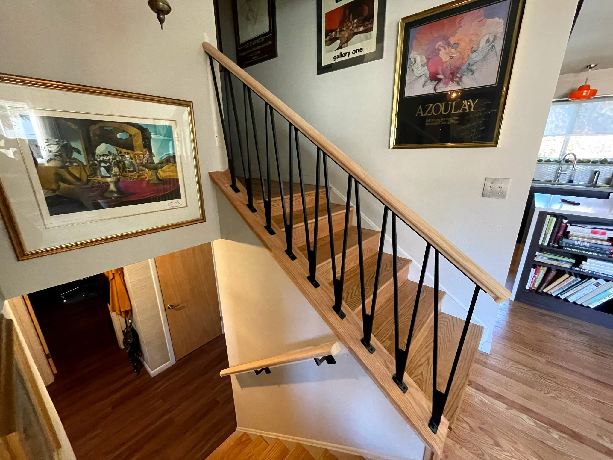 Interior view of a house showcasing a staircase with wooden steps and black metal railings, framed artwork on the walls, and a visible kitchen area with a window and shelves.