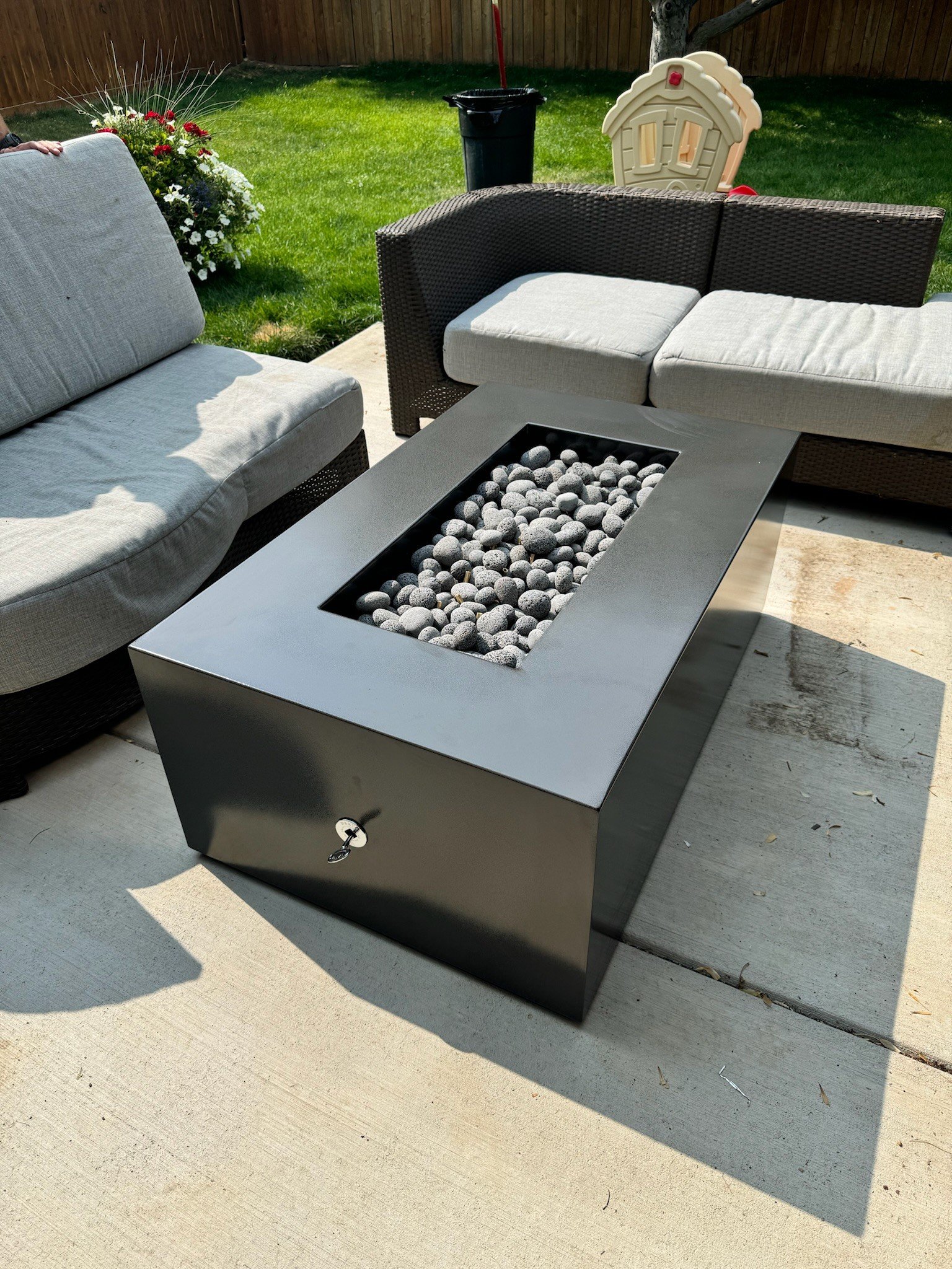Outdoor patio with modern fire pit filled with gray smooth stones, surrounded by cushioned outdoor sofas on a concrete surface, with a grassy yard, potted flowers, a black trash can, and a small plastic playhouse in the background.