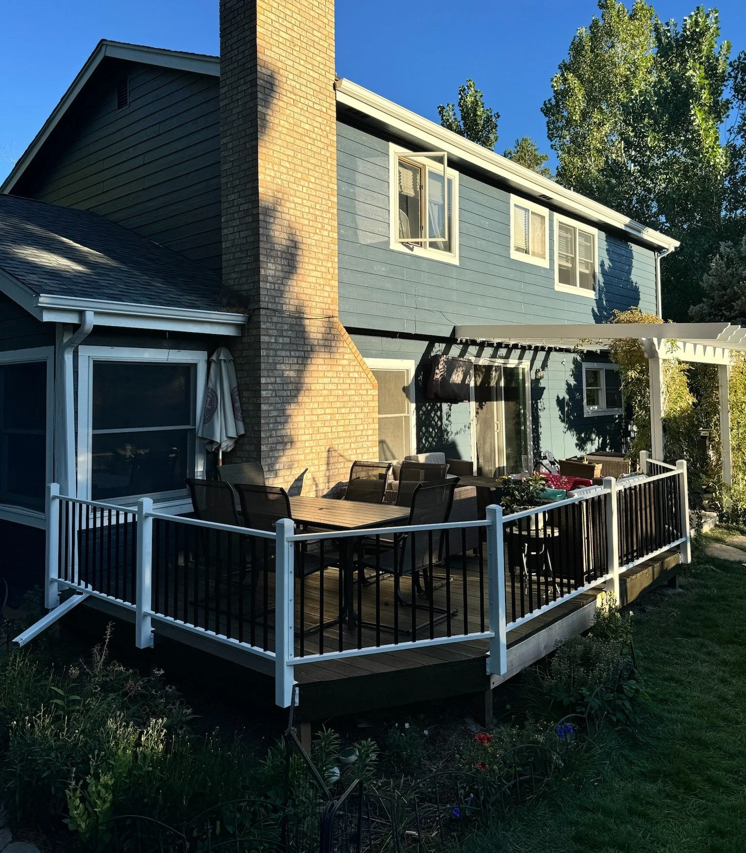 Backyard patio with outdoor dining table, chairs, and a pergola attached to a blue house with multiple windows.