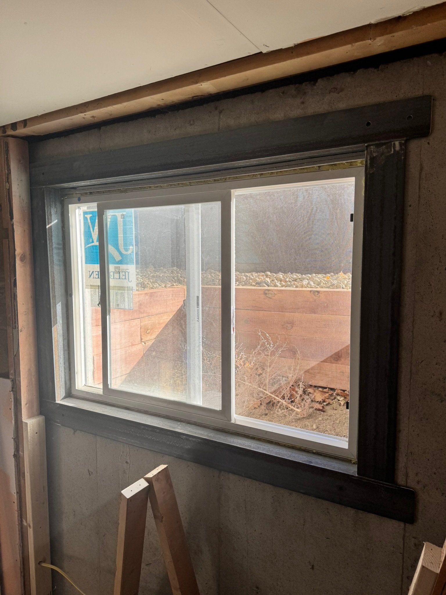 An interior view of a window in a room under construction, with exposed framing and a view of a wooden fence and gravel outside.