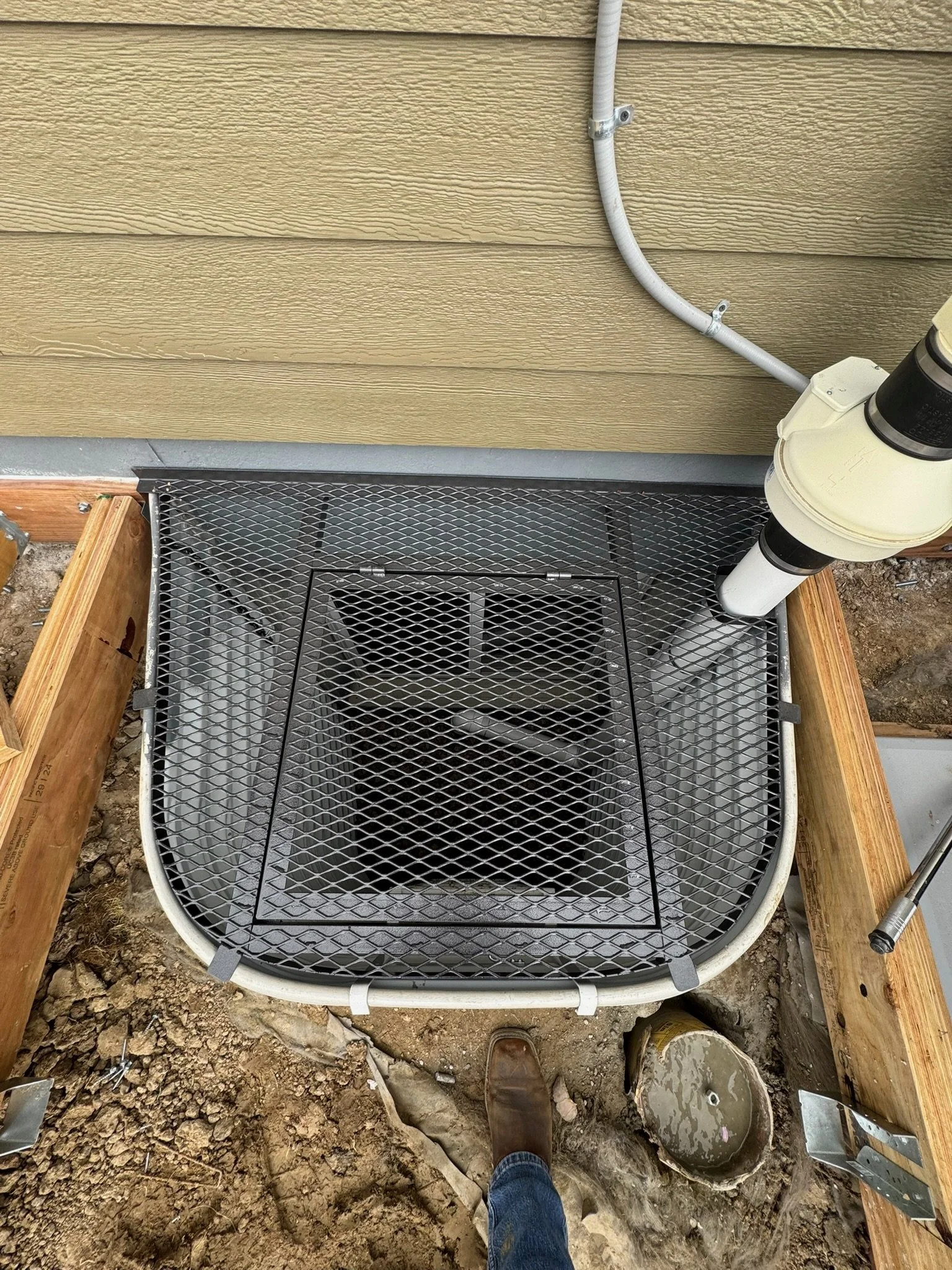 A worker's foot wearing a brown shoe standing on dirt next to a partially completed deck with a metal drain cover, an air conditioning unit, and exterior house siding.