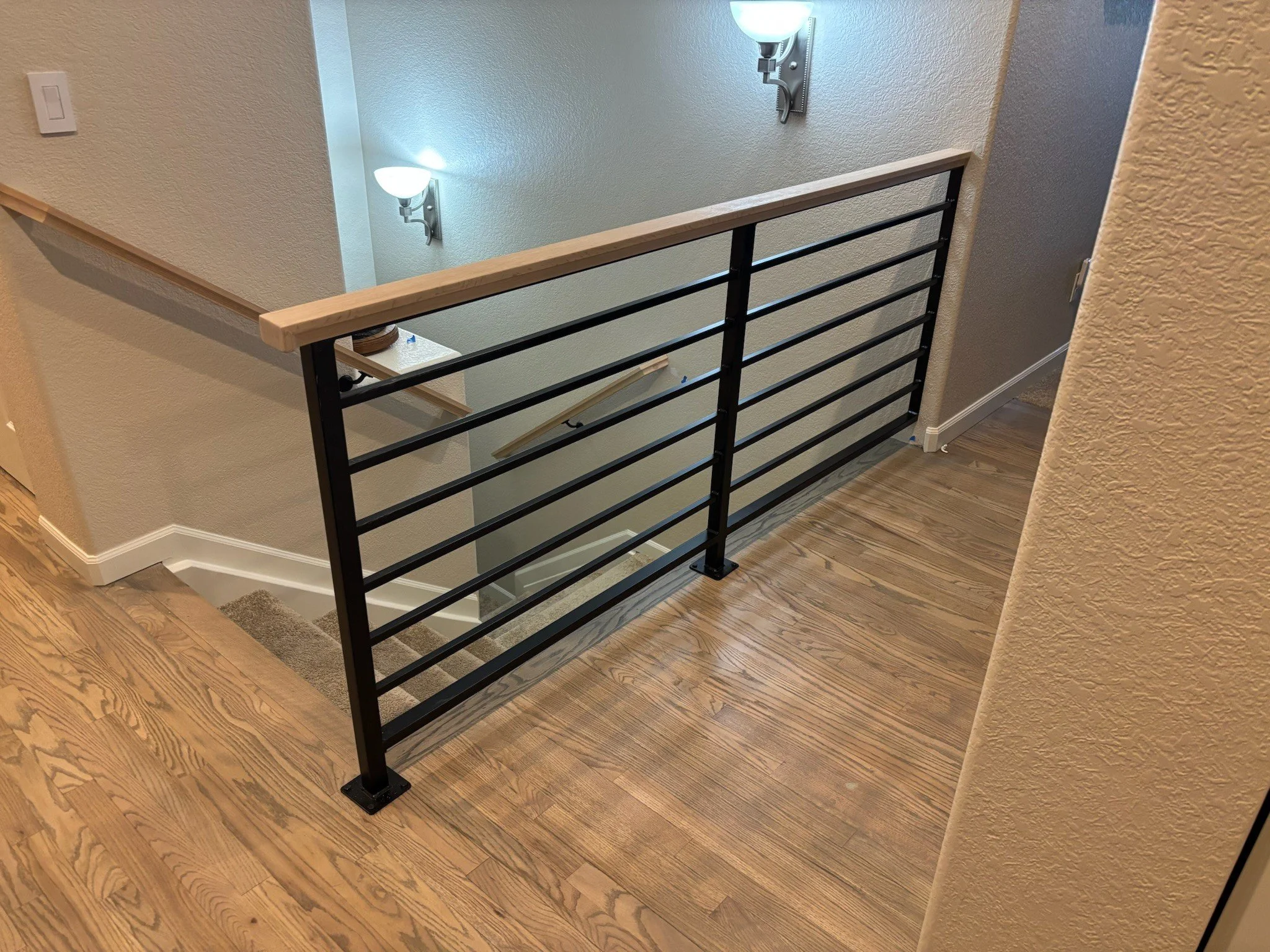Interior view of a second-floor hallway with a black metal railing, wooden handrail, hardwood flooring, textured beige walls, and wall-mounted light fixtures illuminating the space.