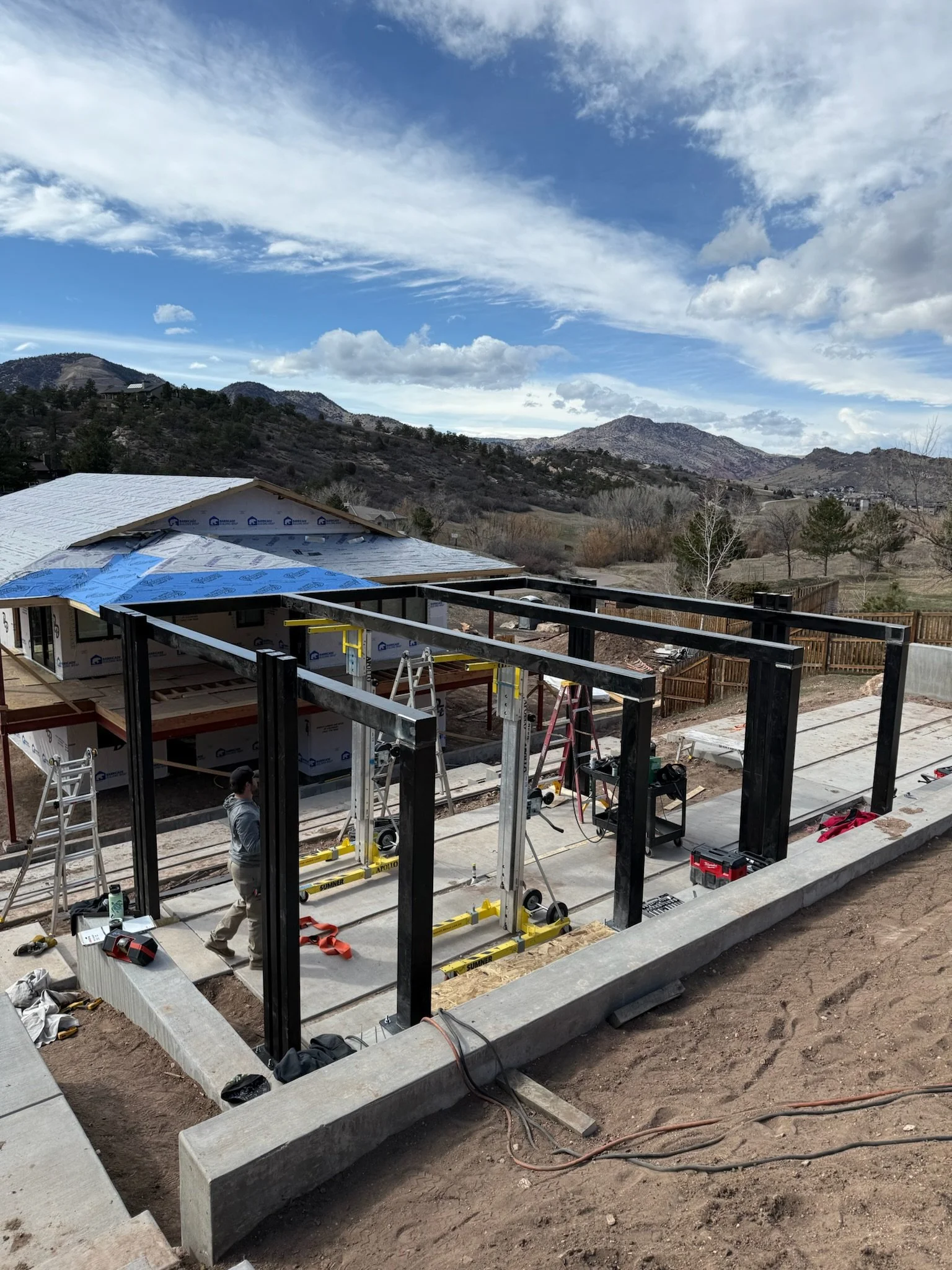 Construction site with black steel framing and concrete slab foundation, in a mountainous rural area under a partly cloudy sky.