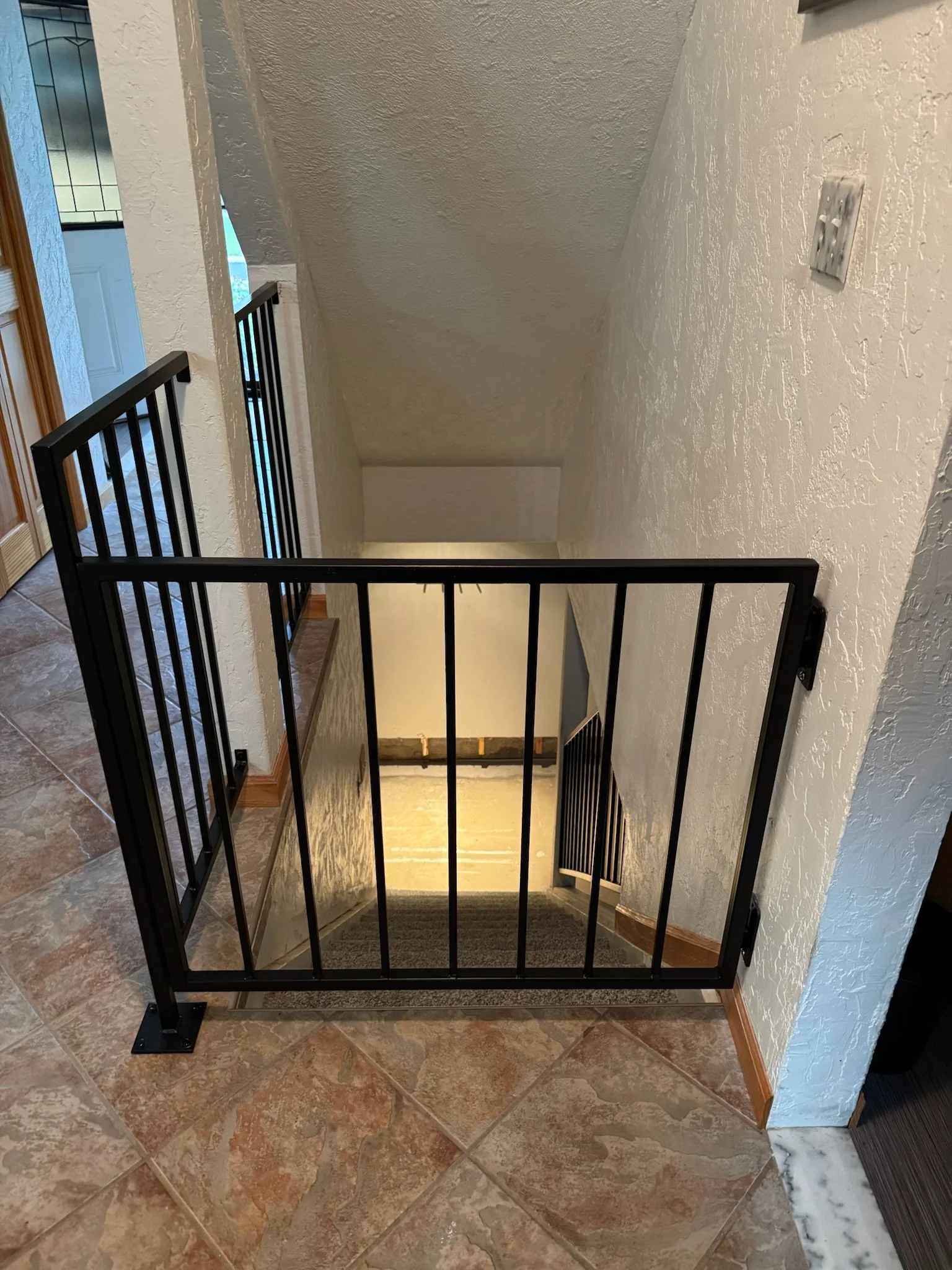 Interior view of a house showing a staircase with a black metal safety gate, beige tiled flooring, and white textured walls.