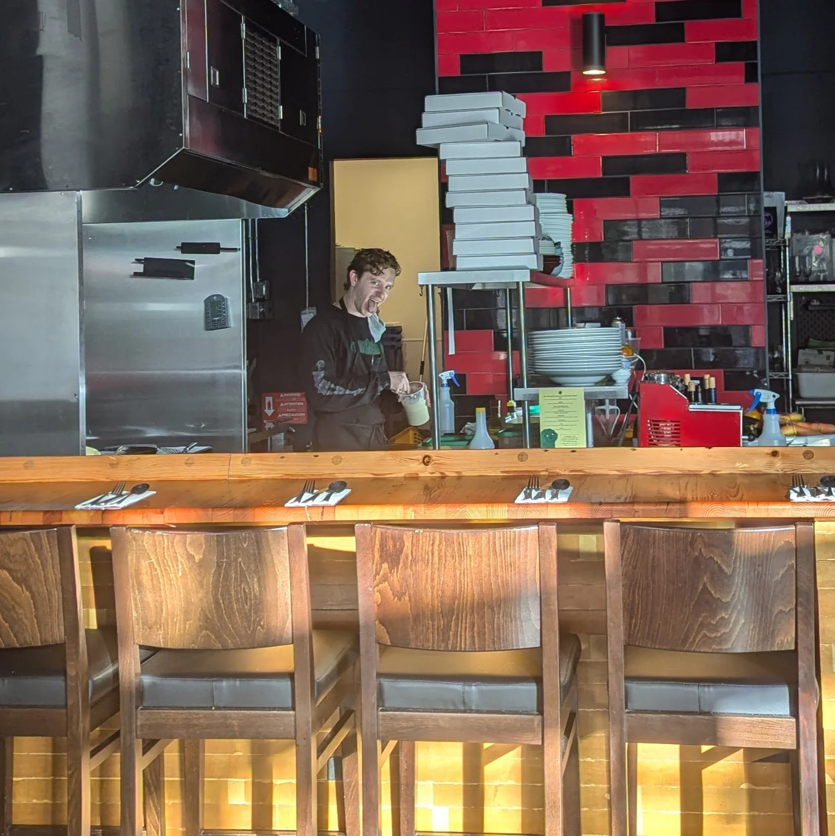A man behind a restaurant counter with stacked boxes and utensils, smiling at the camera.