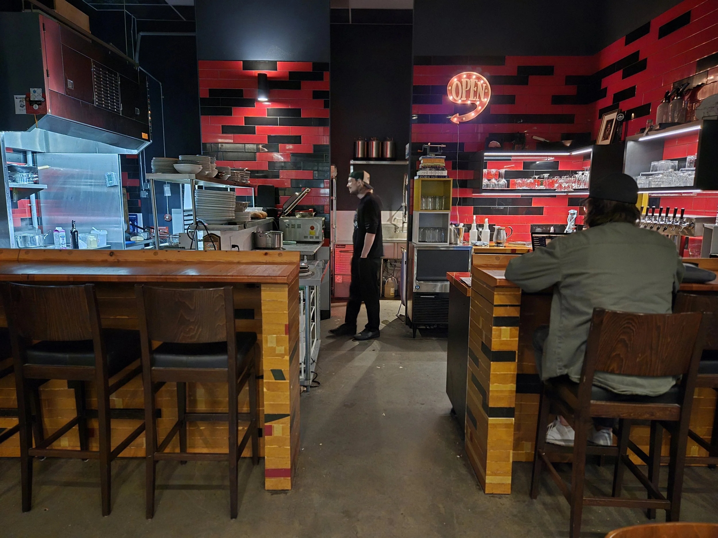 Interior of Hanks a restaurant with a visible kitchen area, a bar counter with a customer sitting, and a staff member walking in the background. Red and black tiled wall with an illuminated "OPEN" sign.