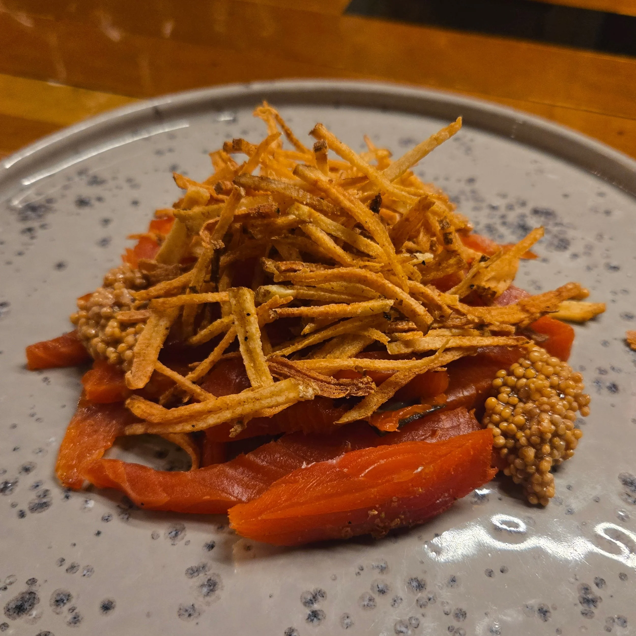 peat smoked sockeye salmon topped with crispy fried potato strings and mustard seeds on a speckled ceramic plate.