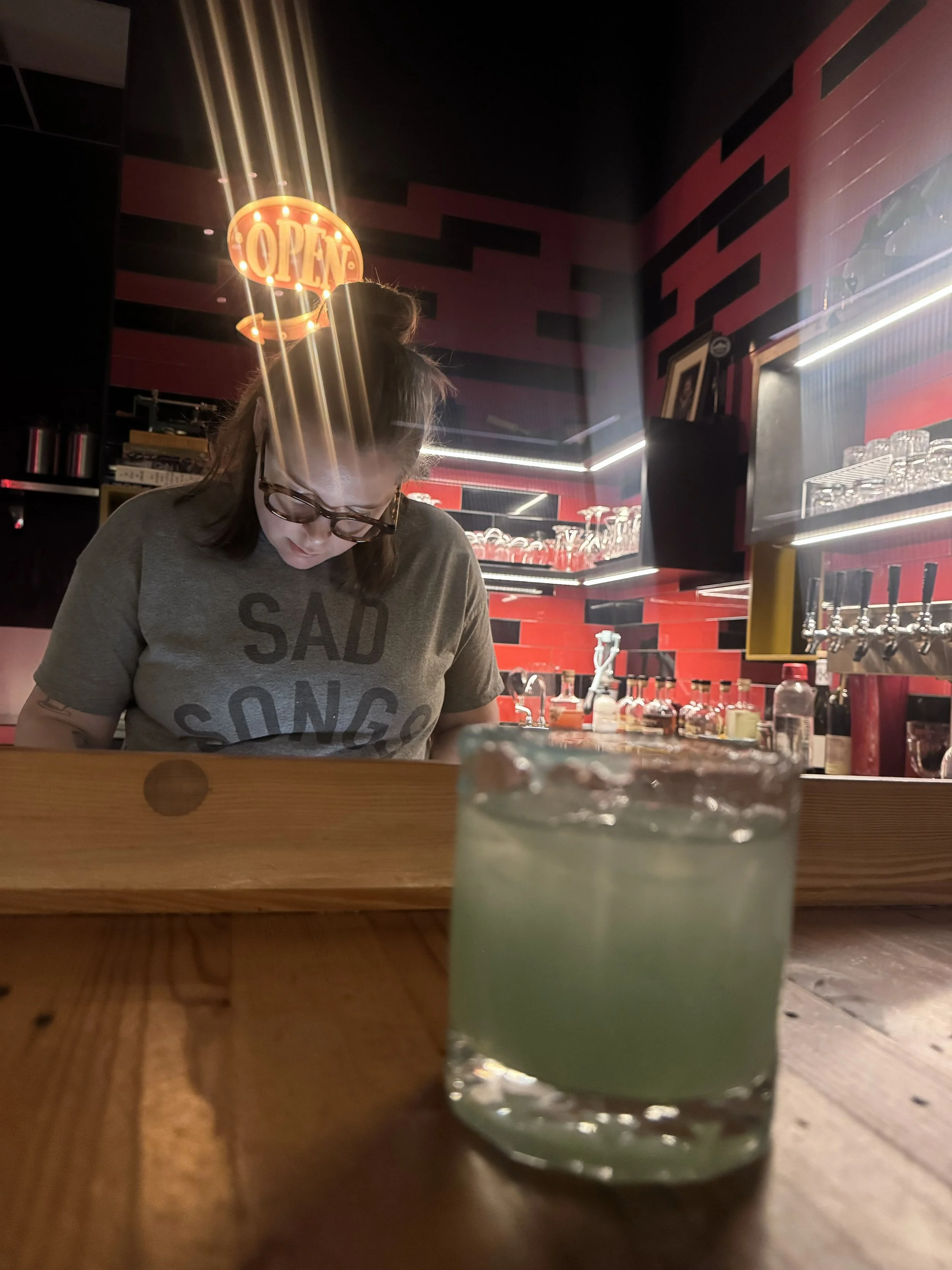 Sara in Hanks a Restaurant with a red and black decor. There is an illuminated "OPEN" sign hanging from the ceiling, and a cocktail with a light-colored beverage and ice sits on the wooden bar in the foreground.