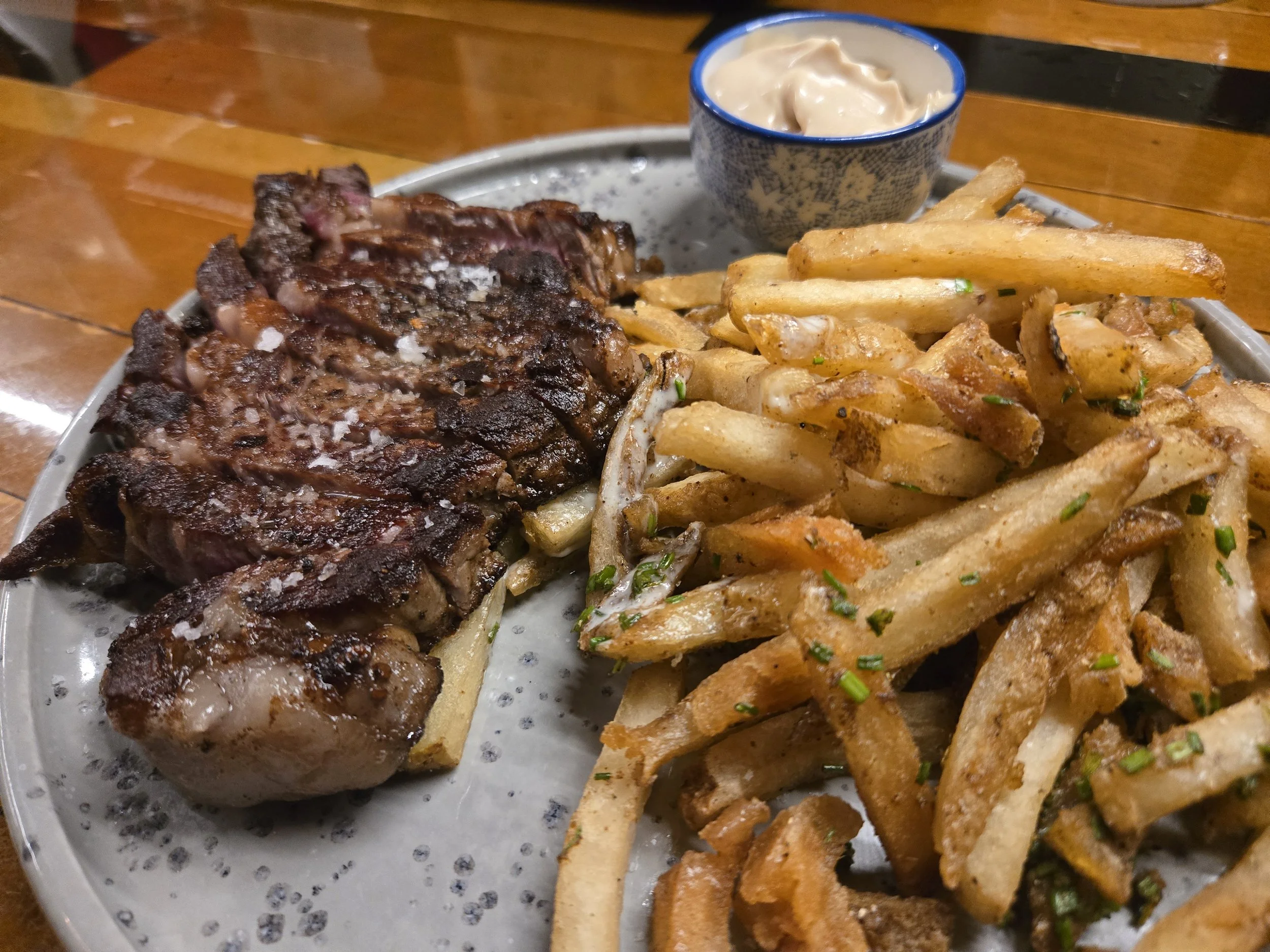 40 day dry-aged ribeye steak, house made French fries topped with chopped herbs, and a small bowl of dipping sauce on a metal tray.