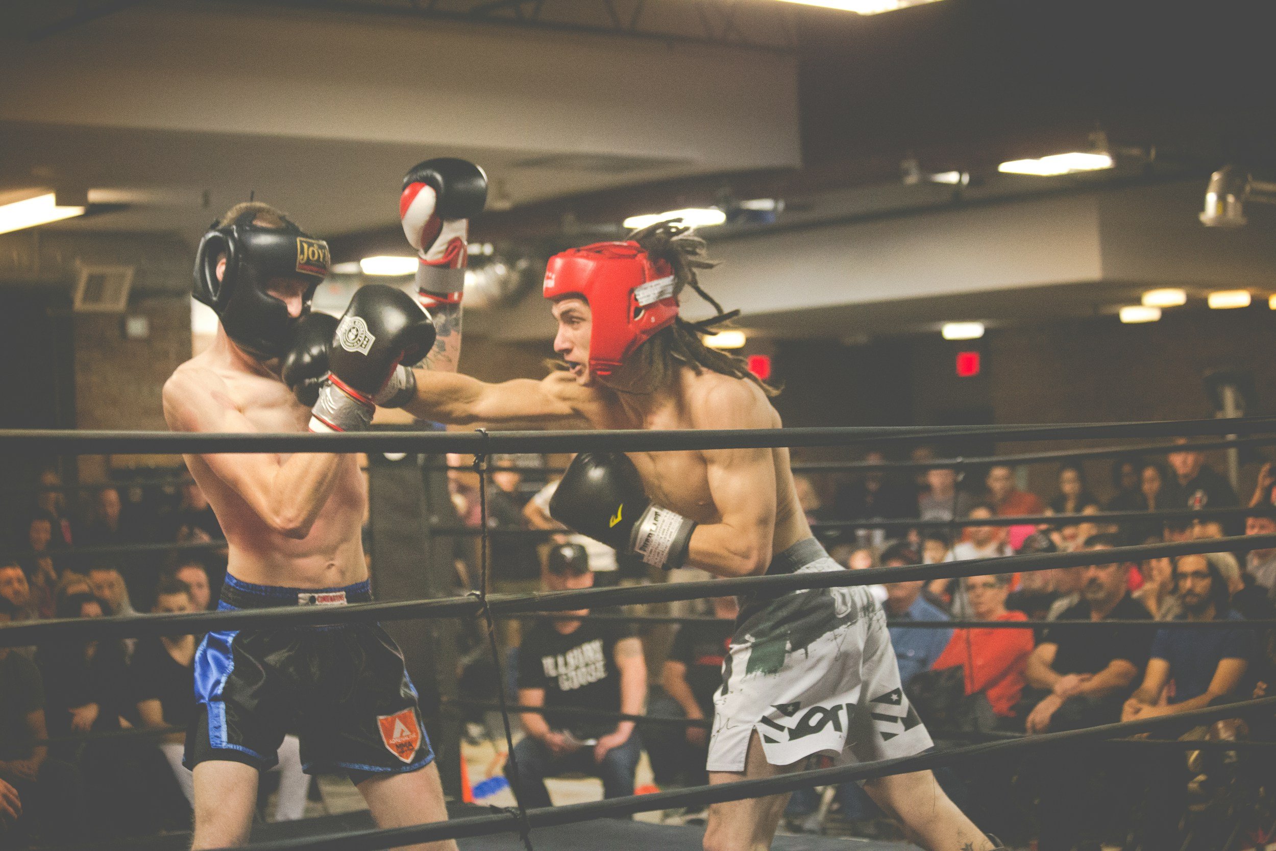 Two male boxers in a boxing ring engaged in a fight, with one delivering a punch to the other's face. Spectators are visible in the background.