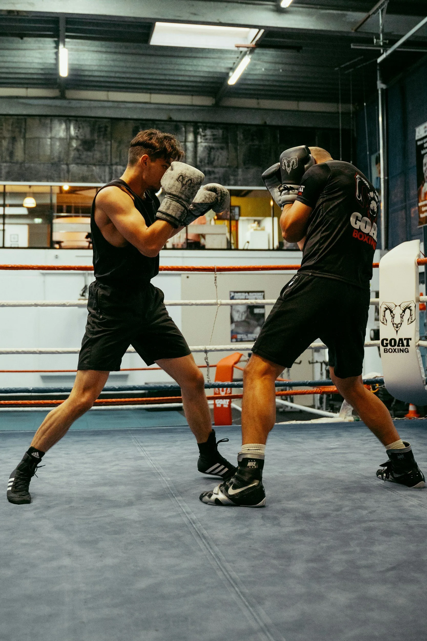 Two young men wearing boxing gloves and athletic clothing training in a boxing gym, sparring in the ring.