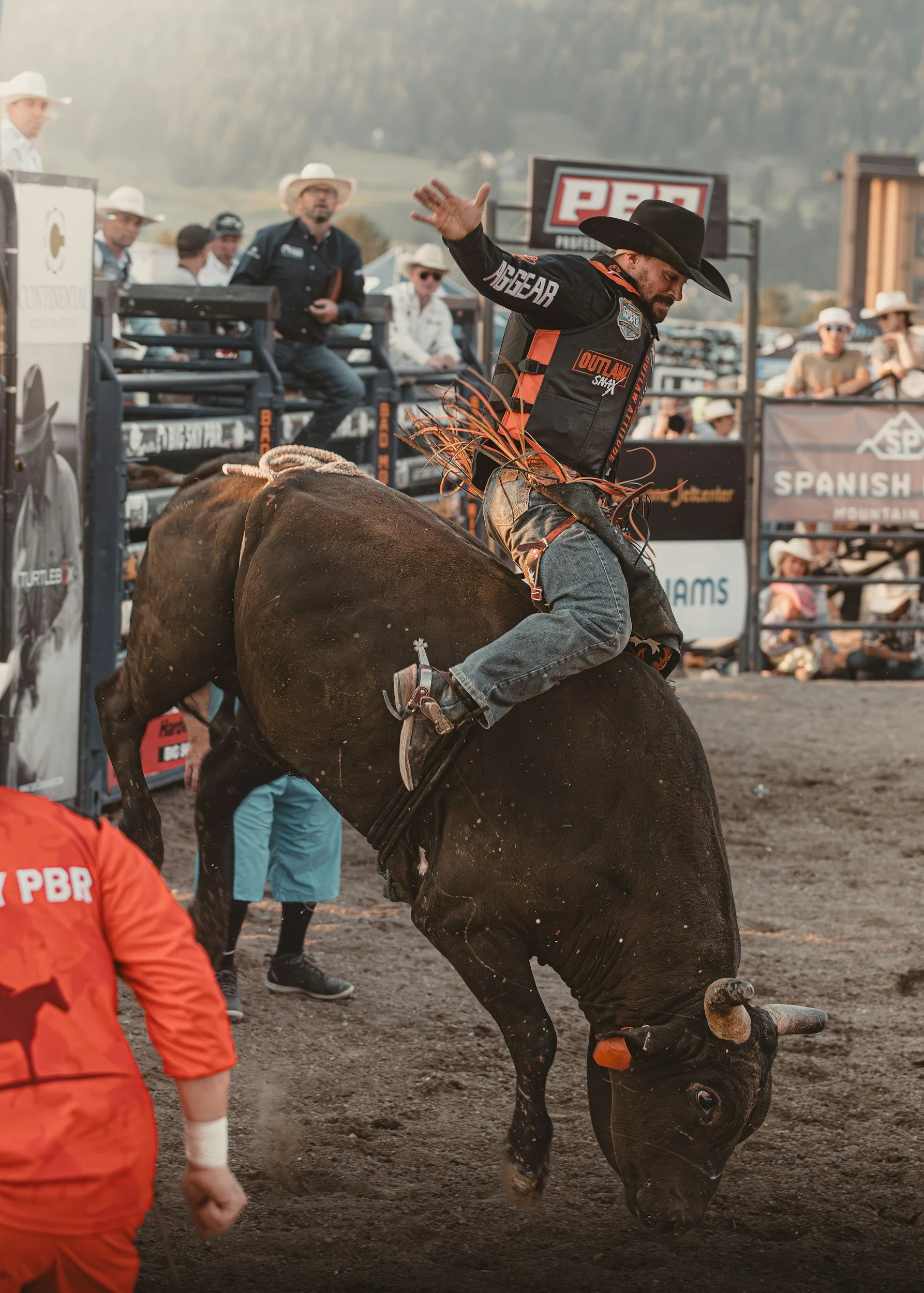 A cowboy wearing a black hat, black shirt, and jeans riding a bucking bull with a brown and black coat, on a dirt rodeo arena with spectators and officials in the background.