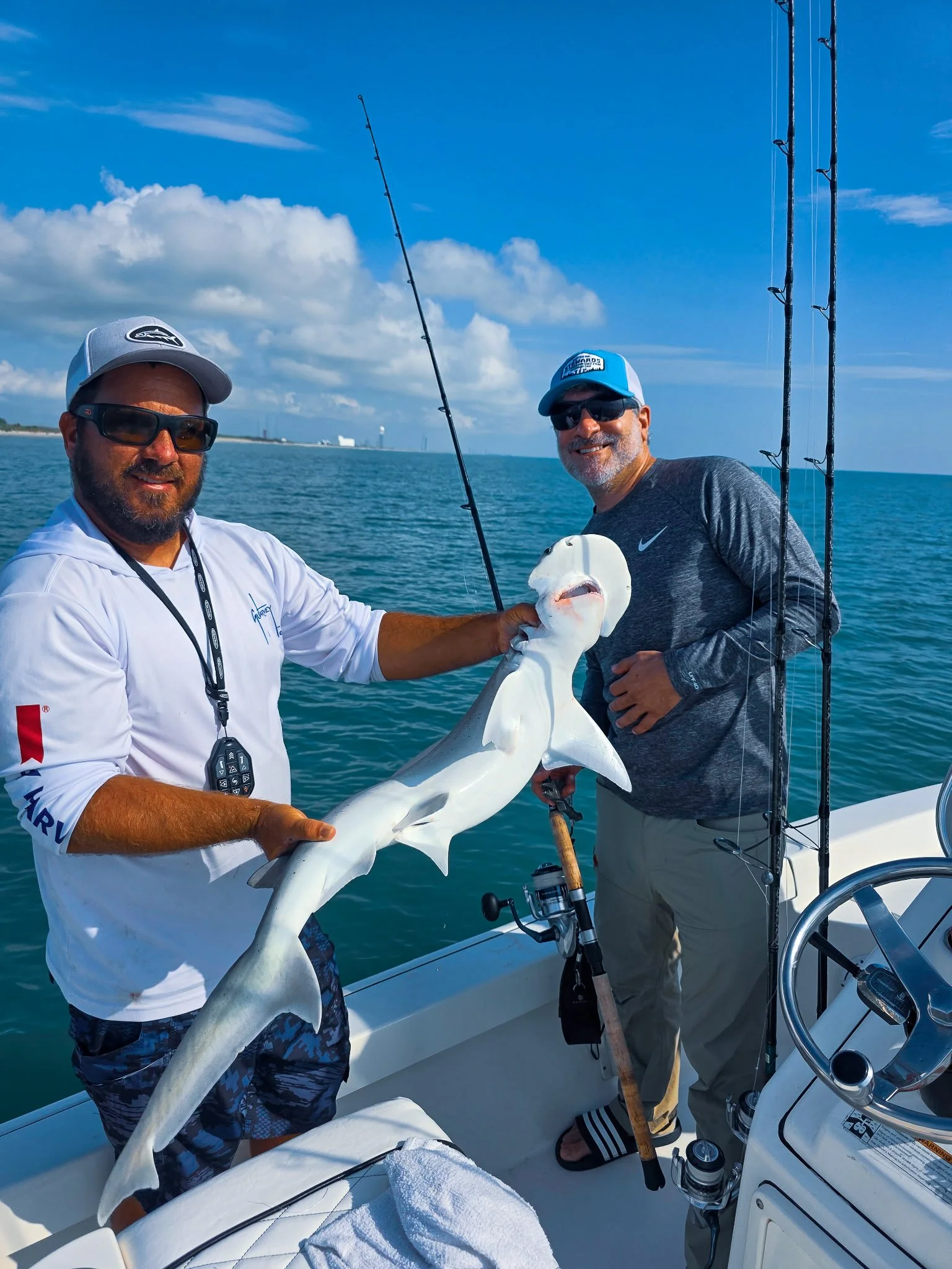Two men on a boat holding a large squid, with fishing rods visible in the background, on a sunny day with blue sky and water. ICast Fishing trip.
