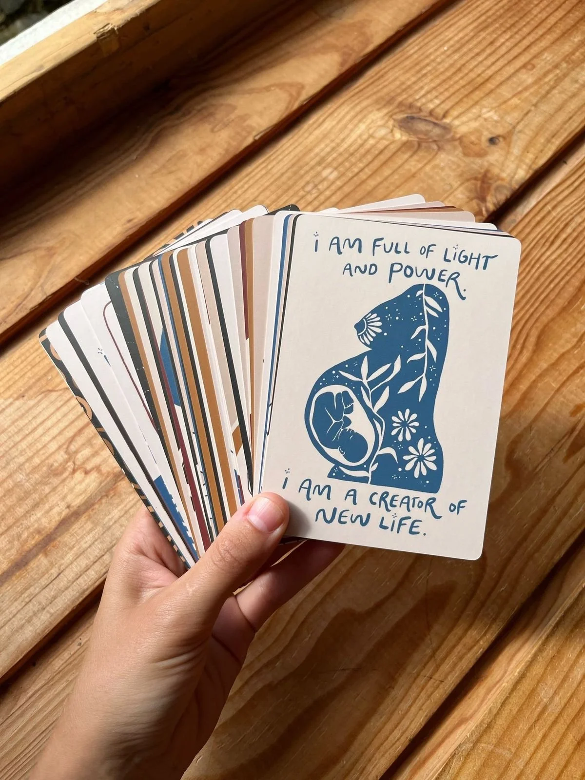 A person holding a deck of cards with one card visible, featuring a blue illustration of a woman with floral and leafy designs and a fetus inside her stomach. The card contains the text: "I am full of light and power. I am a creator of new life." The background shows a wooden surface.