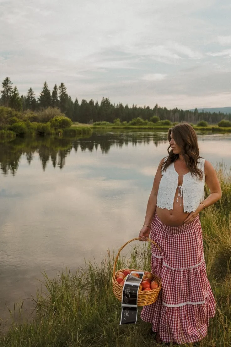 A pregnant woman standing on the grassy bank of a calm river, holding a basket of apples, with trees and mountains in the background under a cloudy sky.
