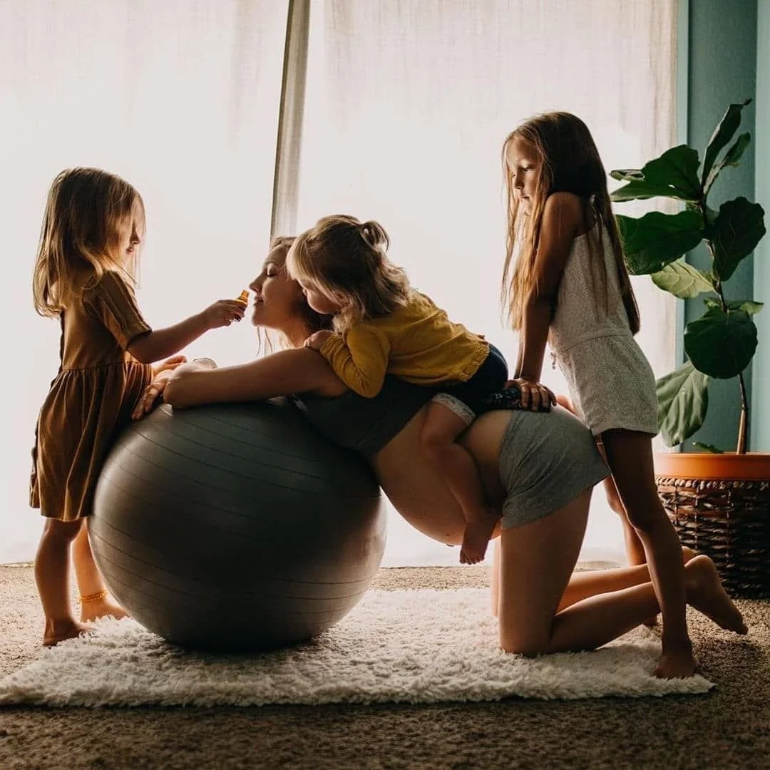 A woman doing a plank exercise with three young girls playing around her in a living room.