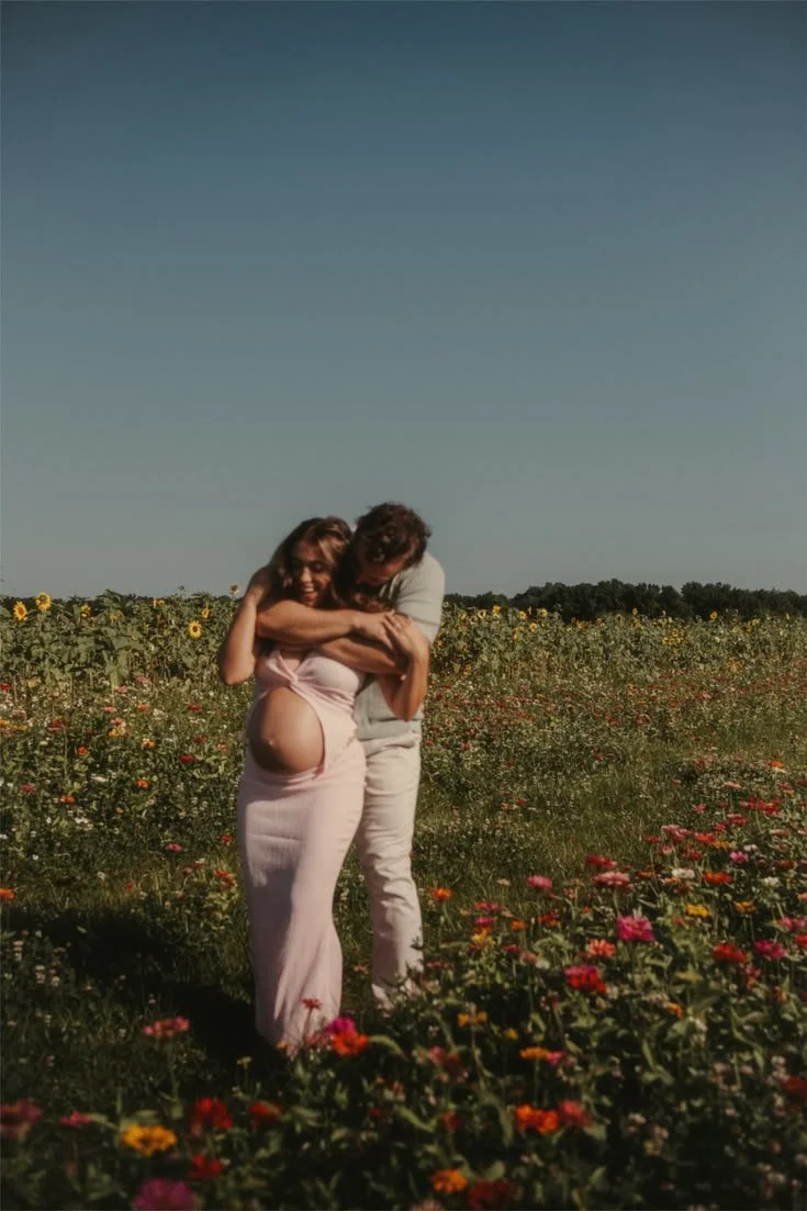 A couple in a flower field, with the woman pregnant, embracing and smiling.