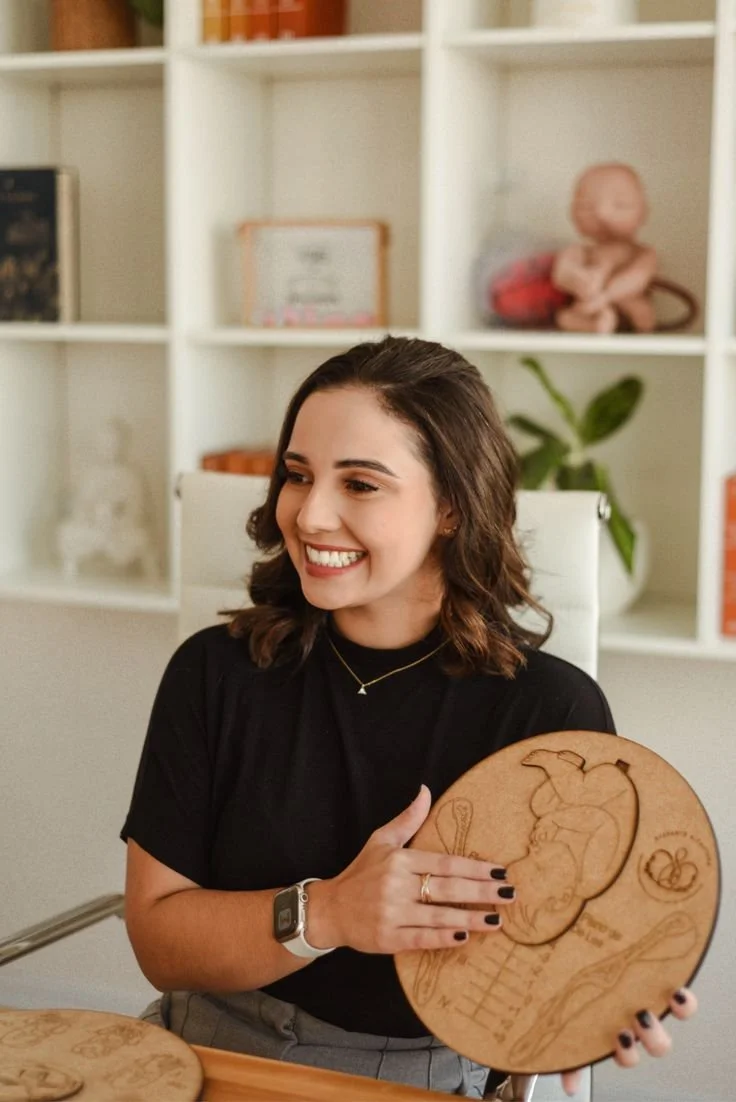 A smiling woman sitting at a desk holding a wooden plaque with engraved artwork of a person in a chef's hat.