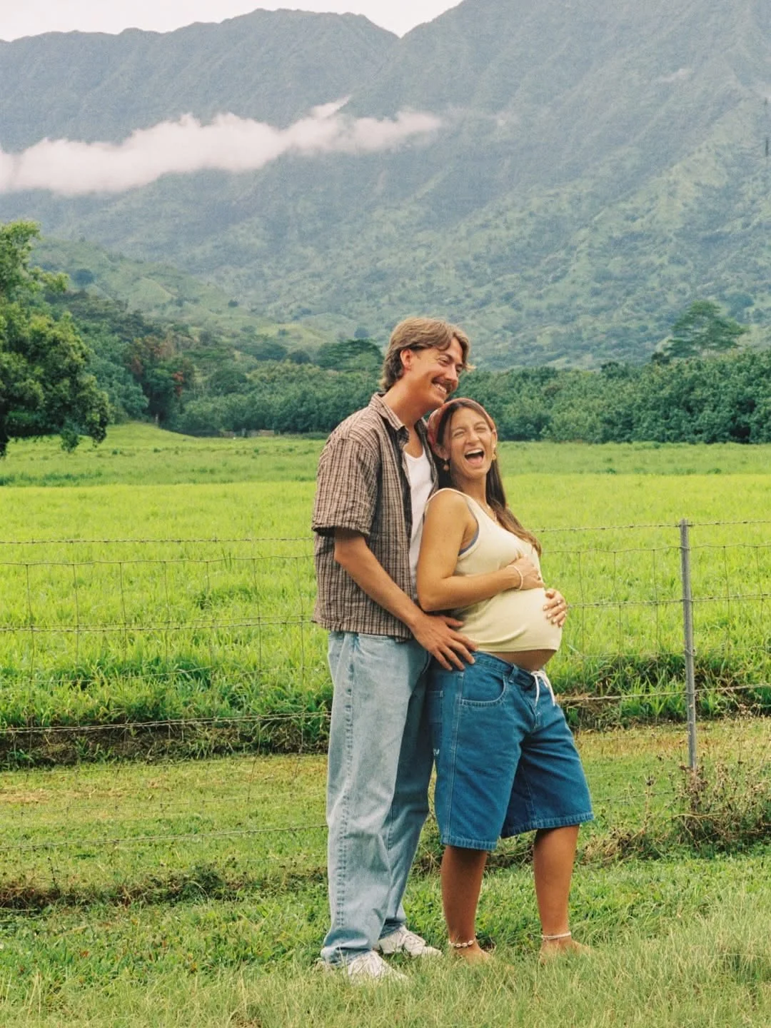 A joyful pregnant woman in casual clothes standing outdoors with a man in a checkered shirt and jeans, both smiling and laughing in a lush green field with mountains in the background.