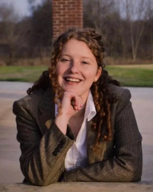 A young woman with curly hair smiling, wearing a gray blazer and white shirt, sitting outdoors at a table with a brick chimney and a grassy area in the background.