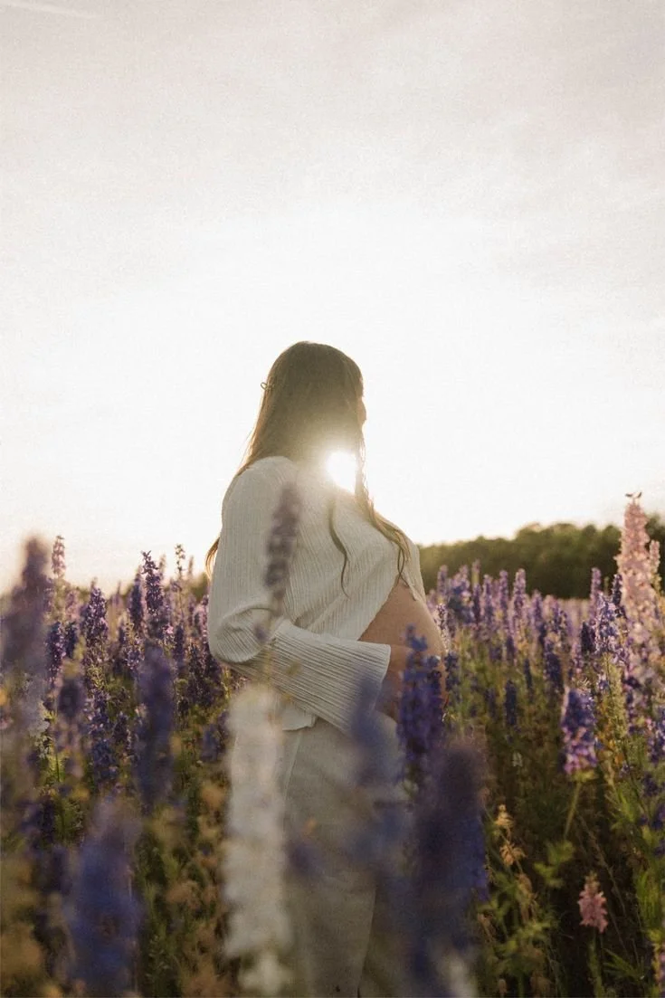 A pregnant woman with long hair and glasses standing in a lavender field at sunset.