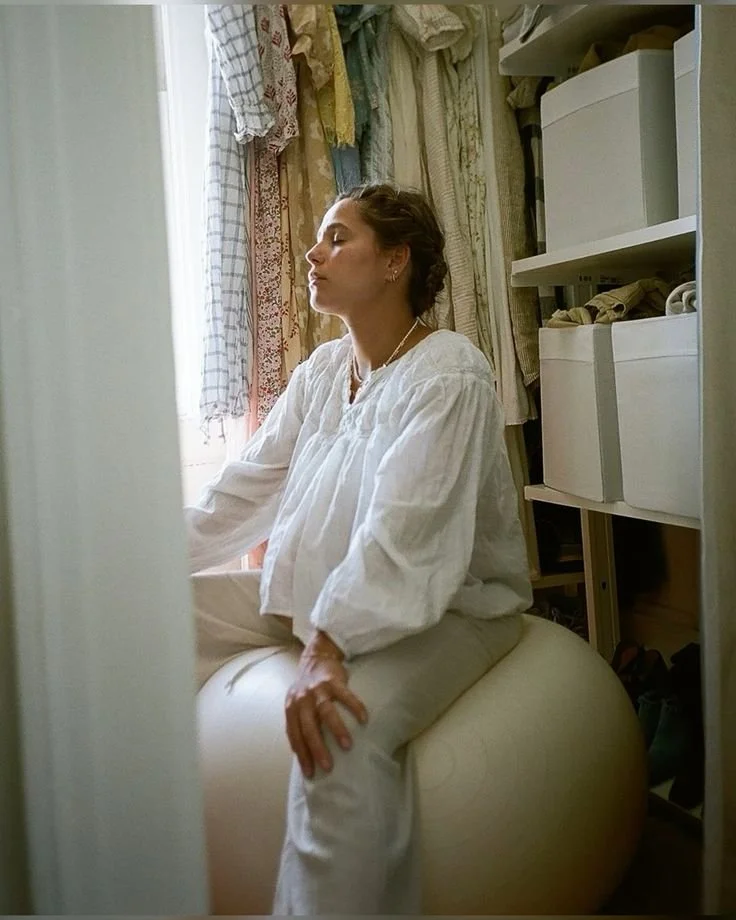 A woman in a white blouse and pants sitting on an exercise ball with her eyes closed in a room with hanging fabric or clothing and shelves in the background.