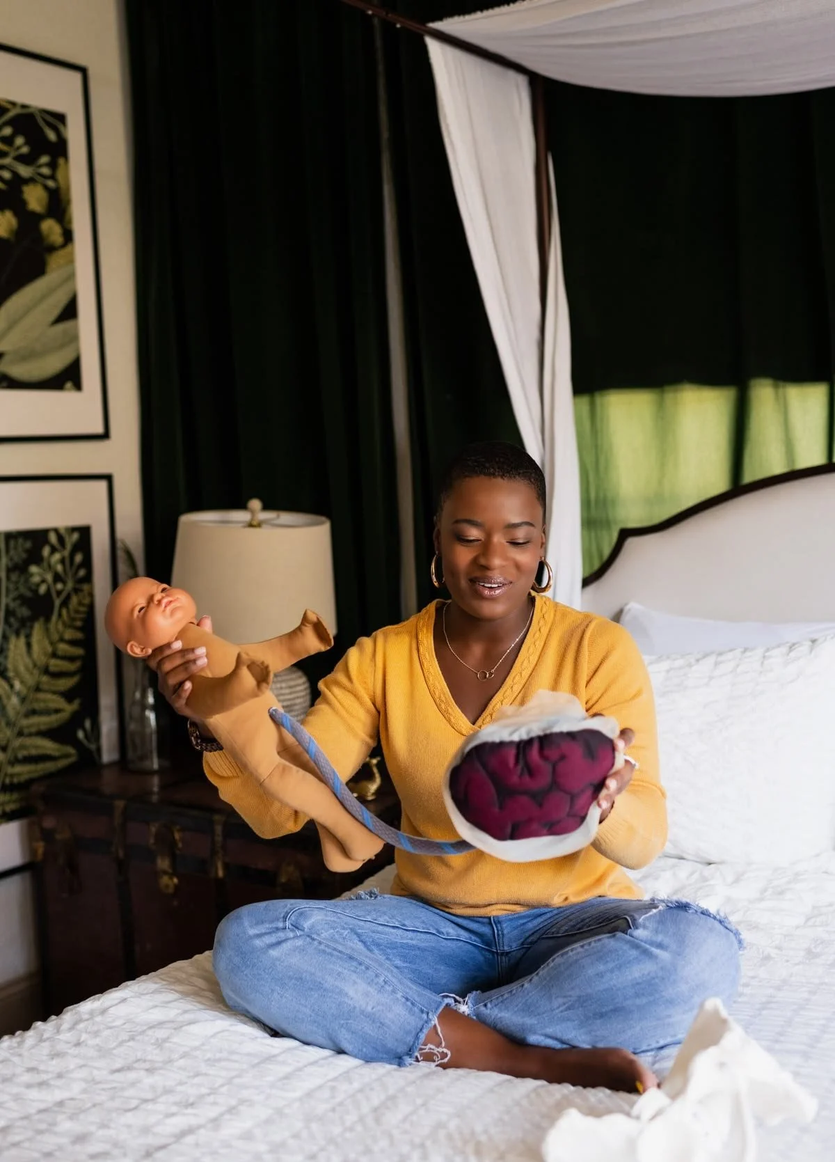 A woman sitting cross-legged on a bed holding a toy brain and a baby doll in a room with dark curtains, a dresser, and a lamp.