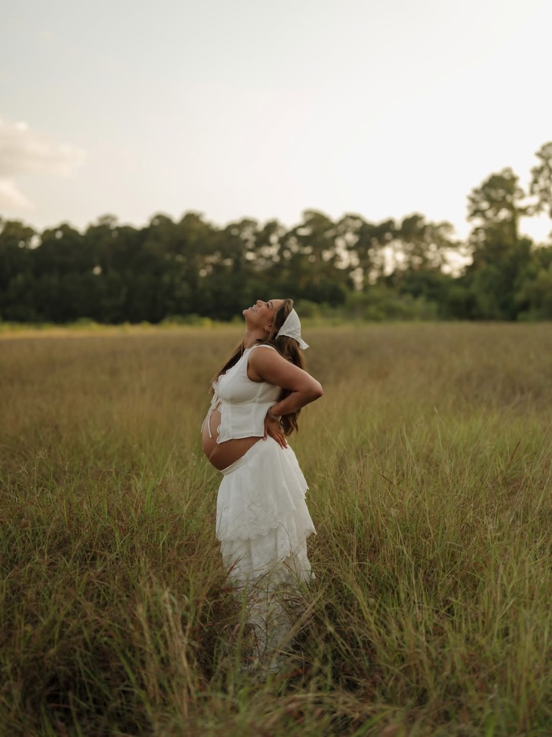 A pregnant woman in a white dress and headscarf standing in a field, smiling and enjoying the outdoors at sunset.