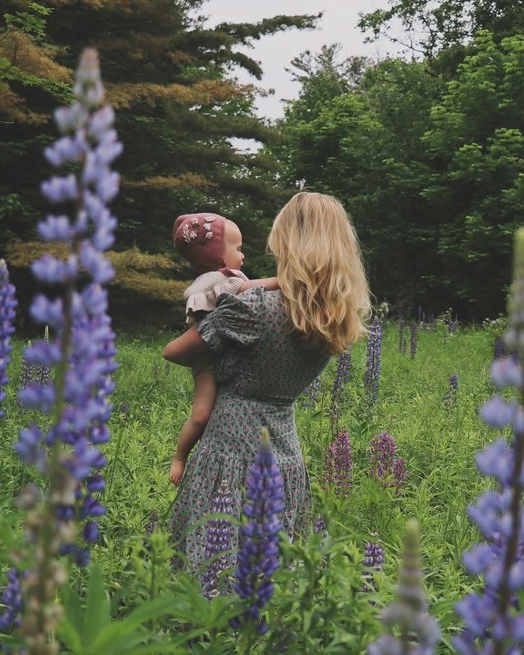 A woman with blonde hair holding a young girl in a field of purple flowers with trees in the background.