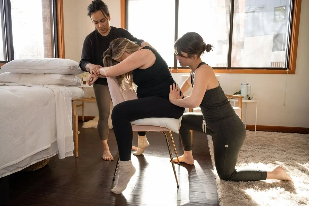 Three women practicing childbirth simulation with one woman seated in a chair, two other women assisting her, in a cozy room with large windows and natural light.