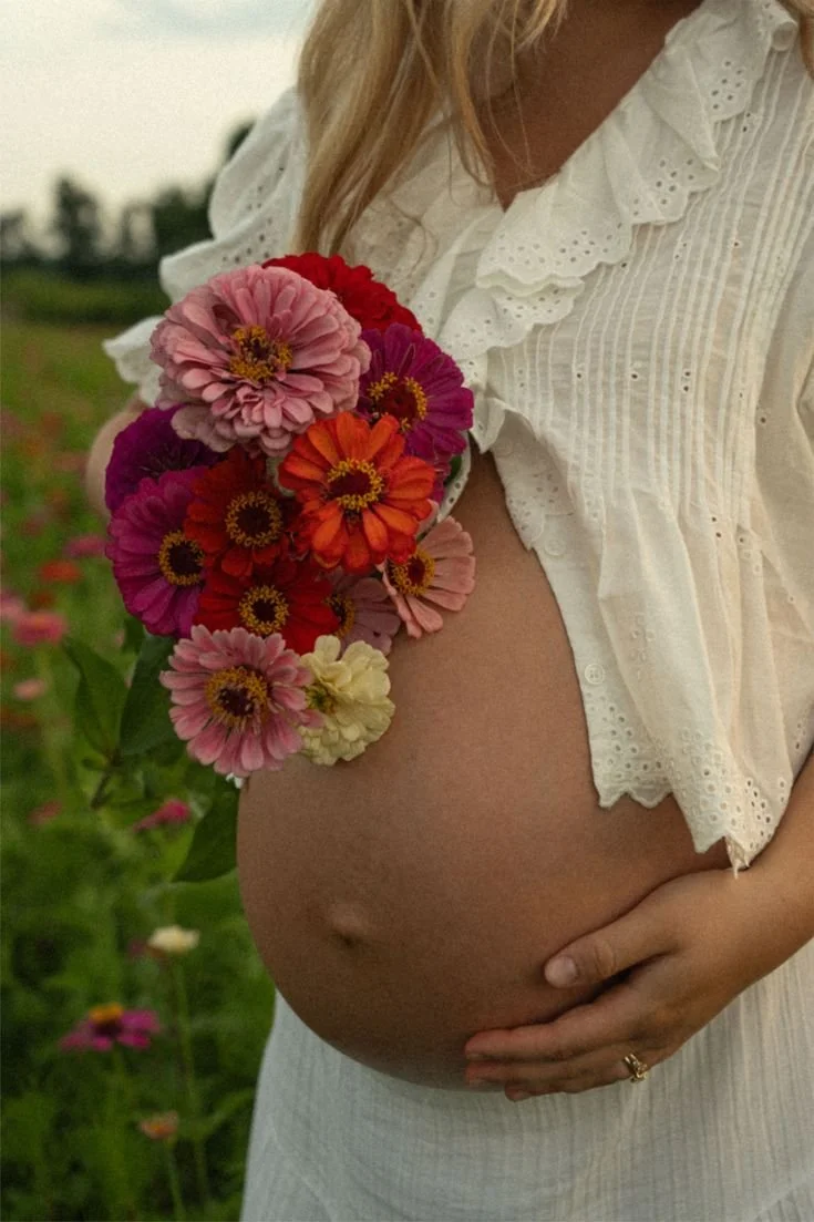A pregnant woman holding colorful flowers in a field, wearing a white blouse and skirt.