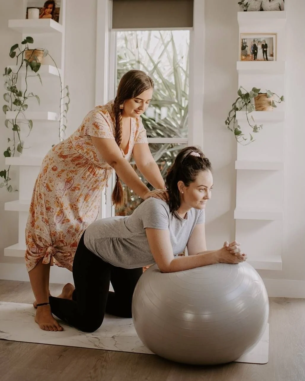 A woman in gray shirt and black pants is doing a plank exercise with her forearms on a large exercise ball, with another woman in a floral dress providing a massage or stretch to her back in a bright, cozy room.