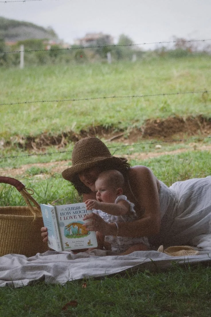 A woman in a wide-brim hat and a light dress lying on a blanket outdoors with a young child, under a tree, reading a child's book titled 'I Guess How Much I Love You,' with a grassy field and fence in the background.