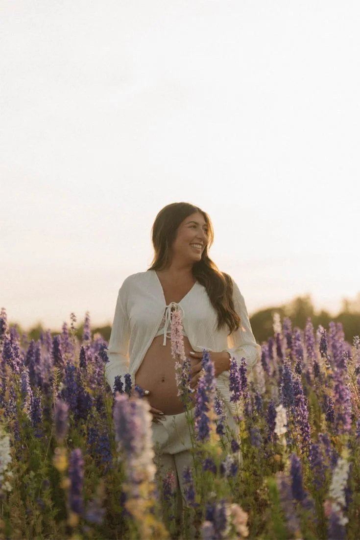 A pregnant woman smiling in a field of purple and pink flowers during sunset.