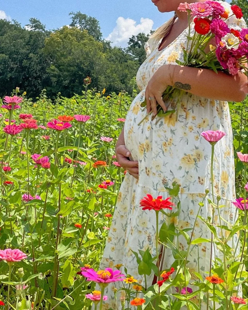Pregnant woman standing in a field of colorful flowers, holding a bouquet of flowers, with trees and a partly cloudy sky in the background.