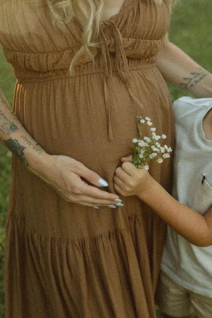 A pregnant woman wearing a brown dress with a drawstring waist, holding her belly with one hand, and a child holding a small bouquet of daisies against her belly.