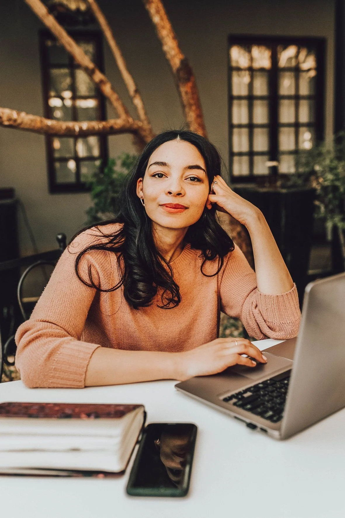 A woman with long black hair, wearing a peach-colored sweater, sitting at a white desk with a laptop, smartphone, and open book, in a cozy indoor space with large windows and a wooden staircase in the background.
