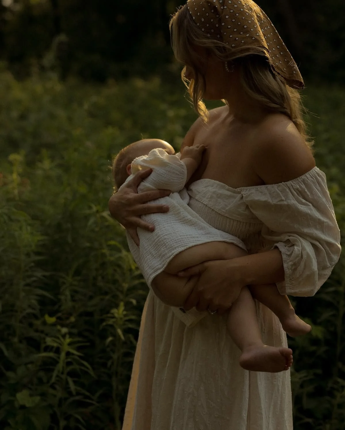 A woman wearing a headscarf and off-shoulder dress holding a baby in a white outfit, breastfeeding outdoors during sunset.