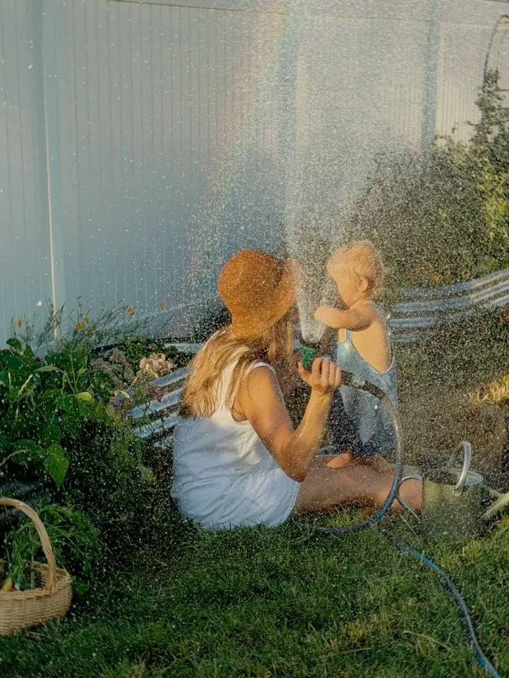 A woman and a young child sitting on the grass while playing with a garden hose, spraying water towards each other during a sunny day.