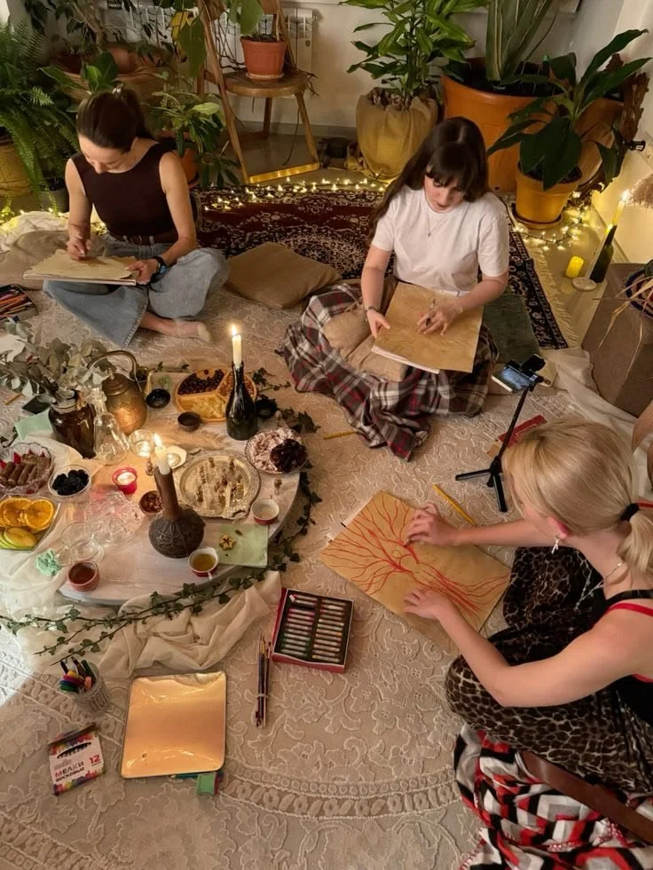 Three women drawing and painting on paper in a cozy room decorated with plants, candles, and snacks.