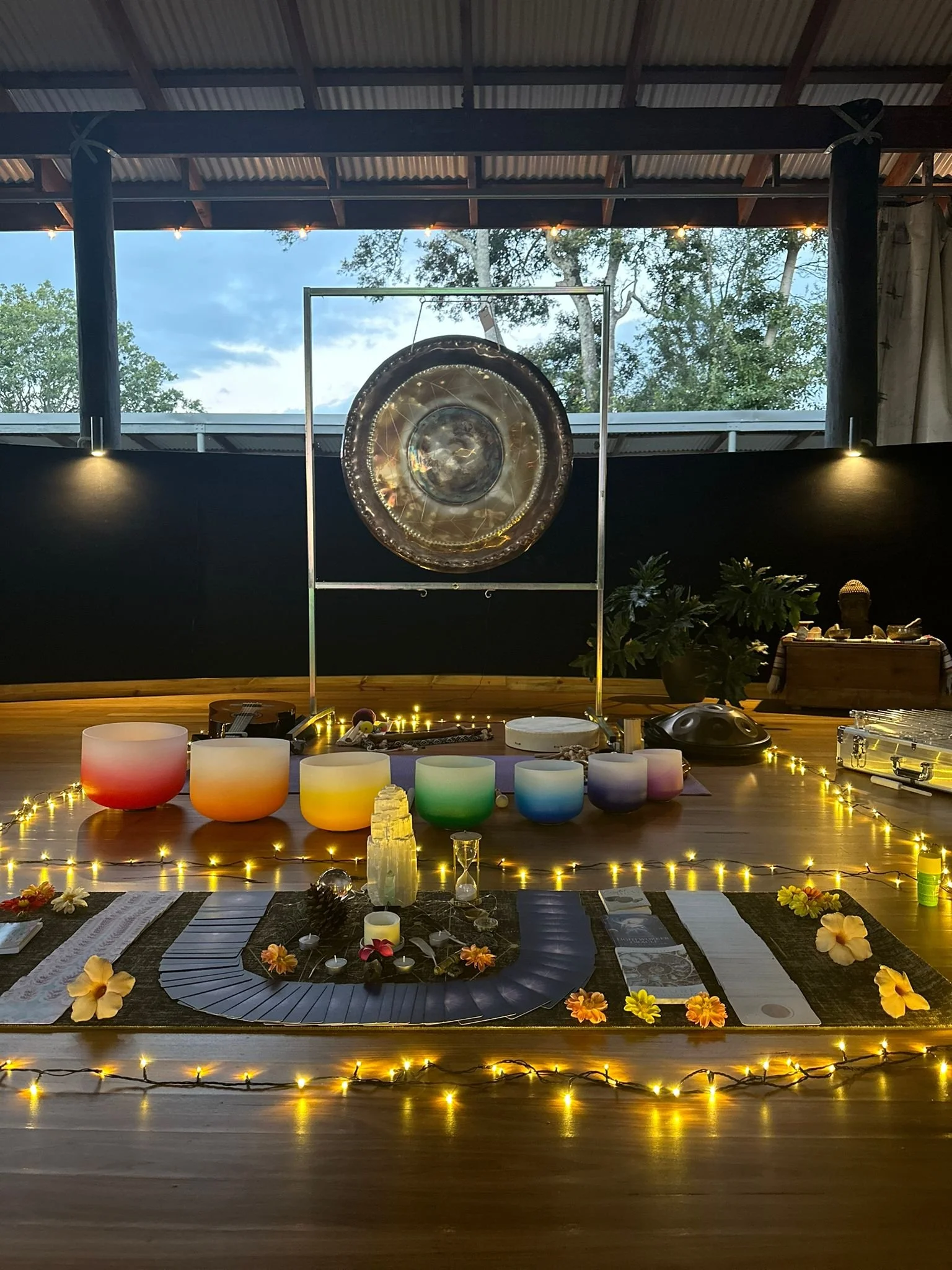 A yoga or meditation setup with colorful singing bowls, candles, flowers, and a gong on a wooden floor, decorated with string lights in an indoor space with large windows showing trees outside.