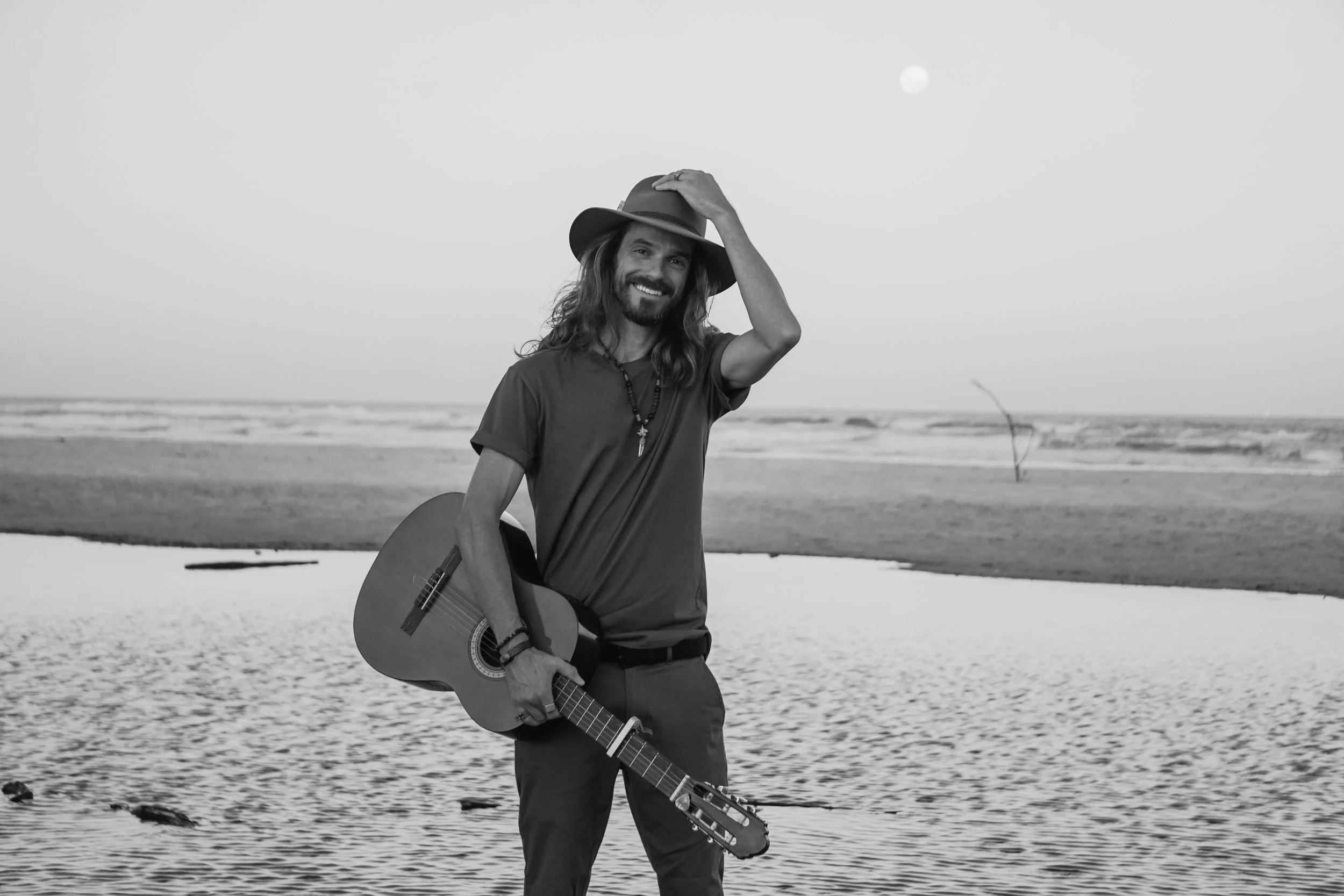 A man with long hair and a beard stands on a beach holding an acoustic guitar. He is smiling and wearing a hat, a casual t-shirt, and necklaces. The background shows sand and water with the moon visible in the sky.