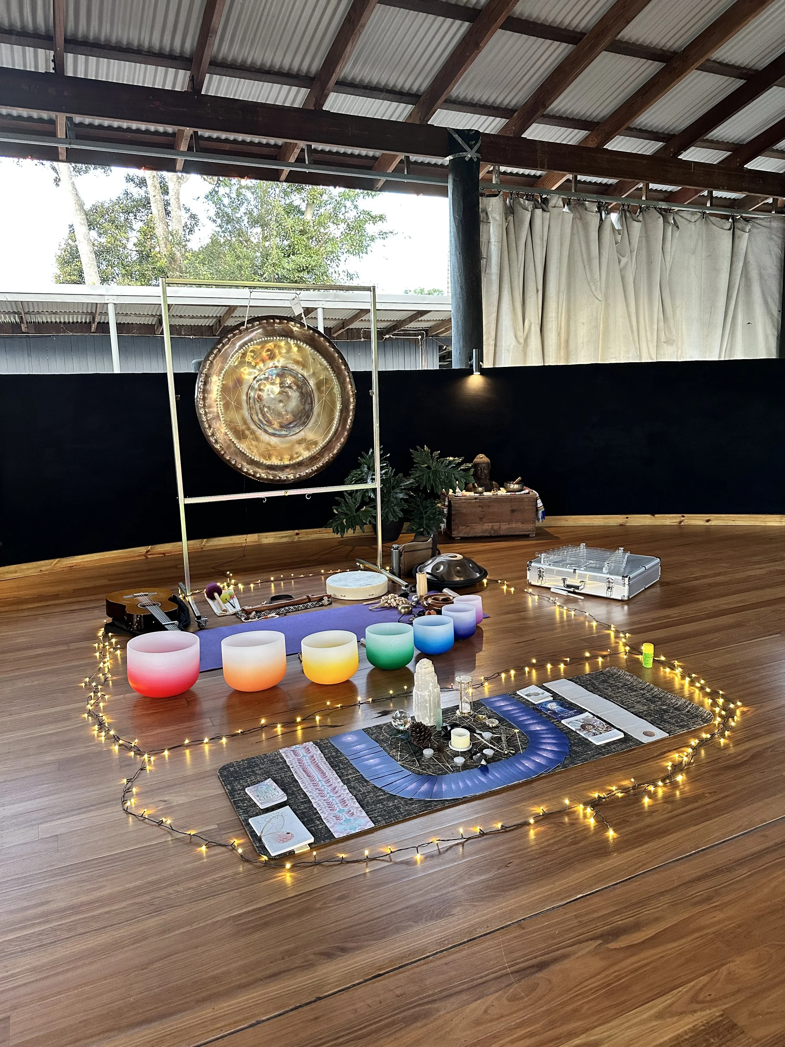 A meditation and sound healing setup on a wooden floor featuring LED candles, crystal singing bowls, tarot cards, a gong, and musical instruments, all surrounded by string lights.