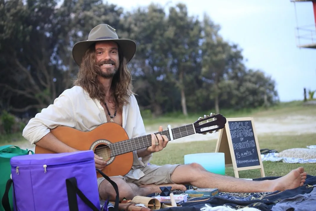 A man with long hair and beard, sitting outside on the grass, playing an acoustic guitar and smiling. He wears a wide-brimmed hat, light-colored shirt, and shorts. There are bags, a sign reading 'Oracle Reading by Donation,' and other items around him.